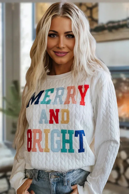Woman wearing a white quilted crewneck sweatshirt with colorful embroidered "MERRY AND BRIGHT" text.