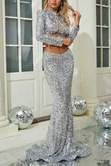 Woman in a silver sequin two-piece evening gown with a crop jacket and mermaid skirt, touching her hair.