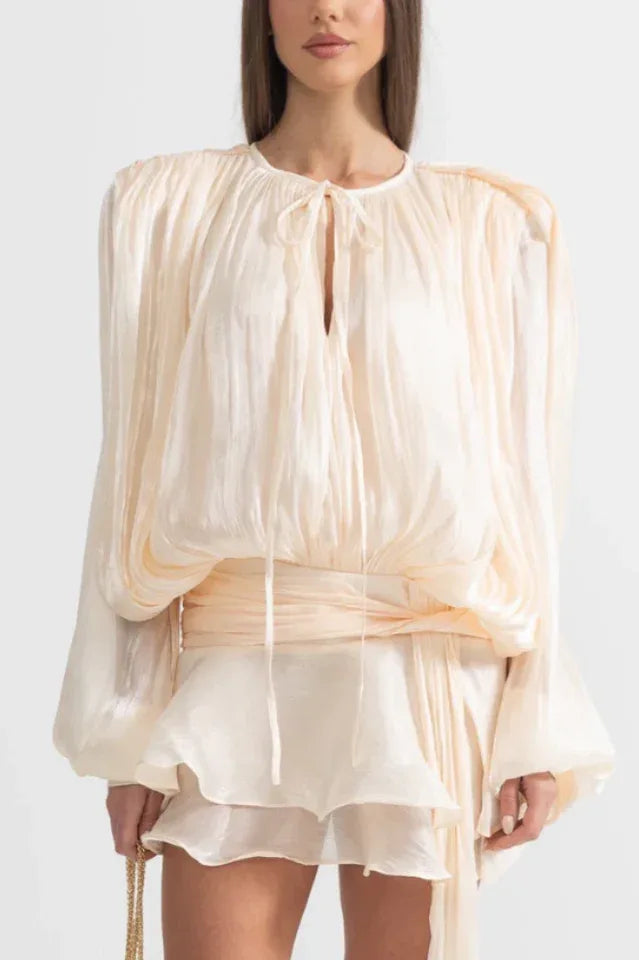 Woman wearing a cream, draped, long-sleeved mini dress with a tie neckline and cinched waist.