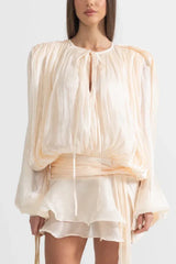 Woman wearing a cream, draped, long-sleeved mini dress with a tie neckline and cinched waist.