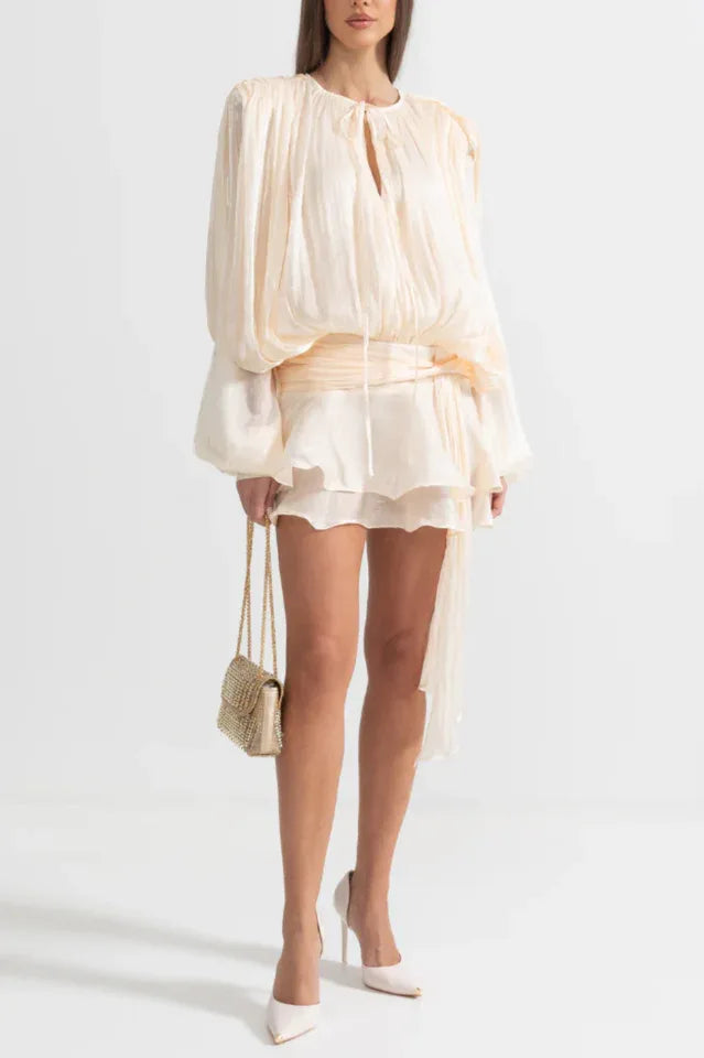 Full-length view of a woman wearing a cream mini dress with balloon sleeves and a cinched waist, holding a gold handbag.