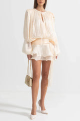 Full-length view of a woman wearing a cream mini dress with balloon sleeves and a cinched waist, holding a gold handbag.