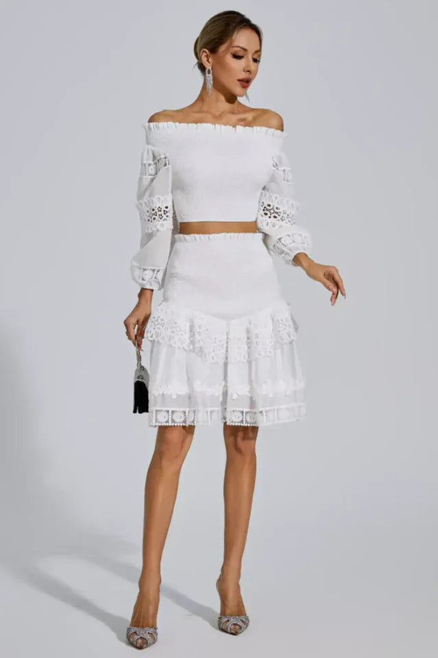 A woman poses in a white smocked crop top and tiered lace skirt, holding a black clutch