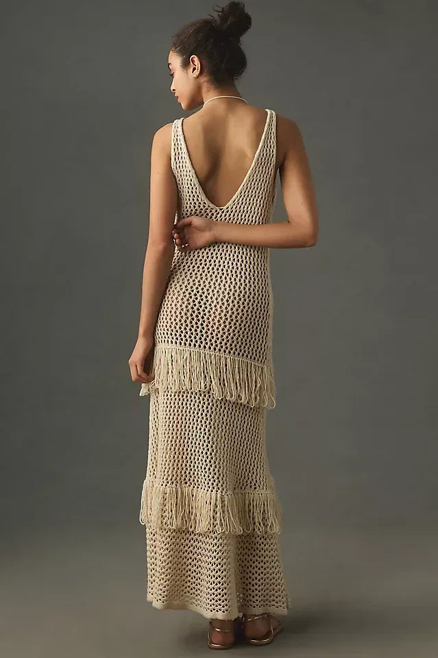 Back view of a woman wearing a cream crochet knit maxi dress with fringe details, standing against a grey background.