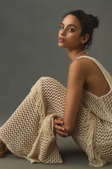 Close-up of a woman wearing a cream crochet knit maxi dress with fringe details, sitting against a grey background.