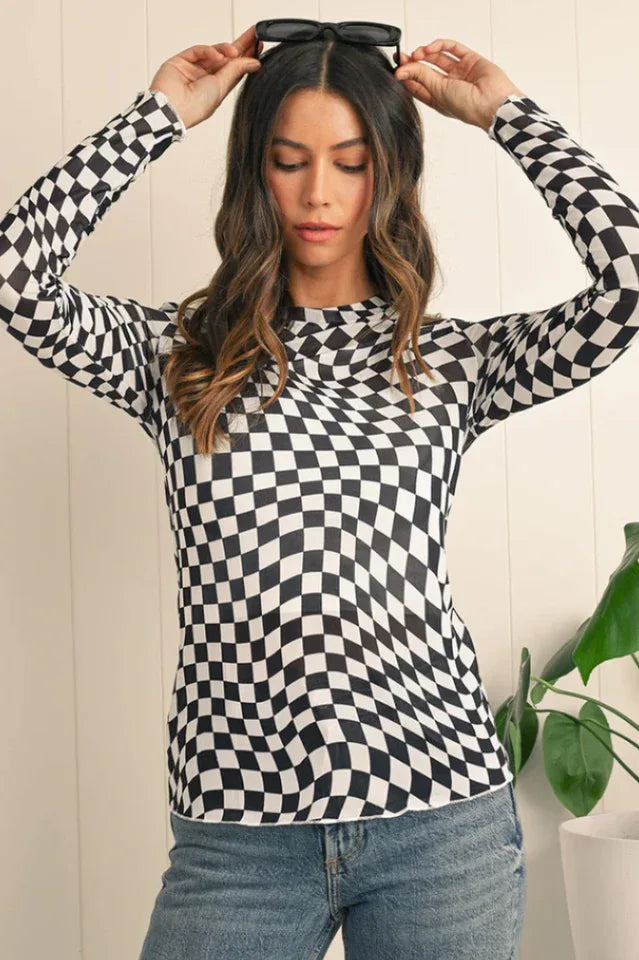 Woman wearing a black and white checkerboard long-sleeved mesh top and blue jeans, holding black sunglasses on her head.