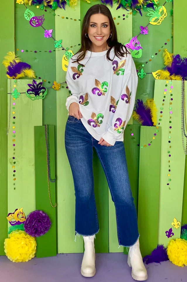 Full body shot of a woman in a white sequin fleur-de-lis sweatshirt and flared jeans, standing in front of a festive Mardi Gras-themed backdrop.