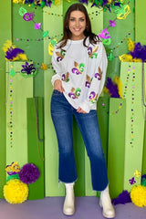 Full body shot of a woman in a white sequin fleur-de-lis sweatshirt and flared jeans, standing in front of a festive Mardi Gras-themed backdrop.