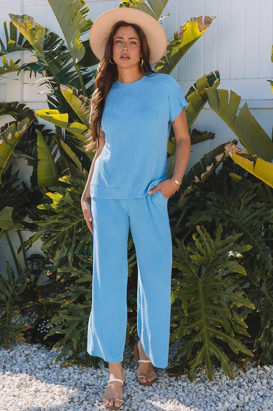 Woman wearing a blue ribbed short-sleeve top and wide-leg pants with a straw hat and sandals, standing outdoors.