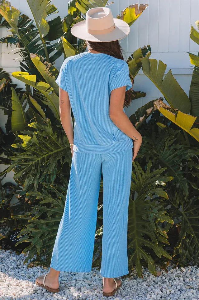 Back view of a woman wearing a blue ribbed short-sleeve top and wide-leg pants with a straw hat and sandals.