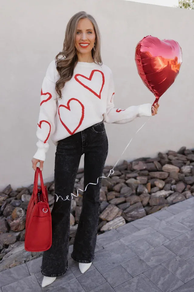 Woman wearing a white knit sweater with a red heart print, black flared jeans, and white boots, holding a red bag and a red heart balloon.