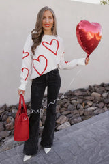 Woman wearing a white knit sweater with a red heart print, black flared jeans, and white boots, holding a red bag and a red heart balloon.