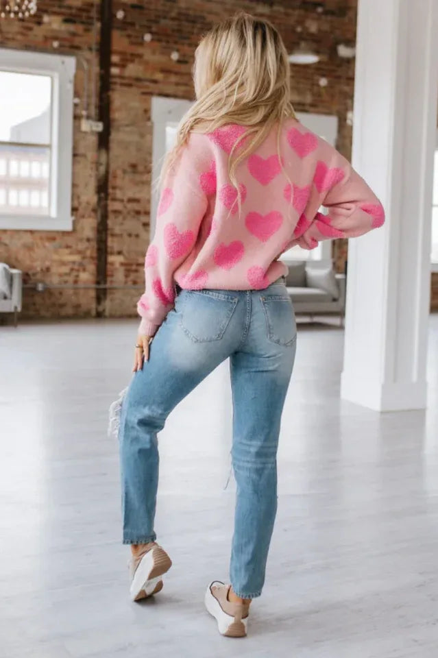 Back view of a woman wearing a pink crew neck sweater with a textured pink heart pattern and distressed blue jeans.