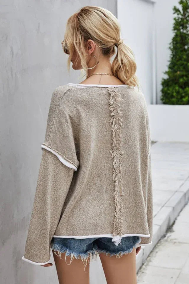 Back view of a woman wearing a beige textured knit sweater with white trim and denim shorts, showing fringe detail on the back.