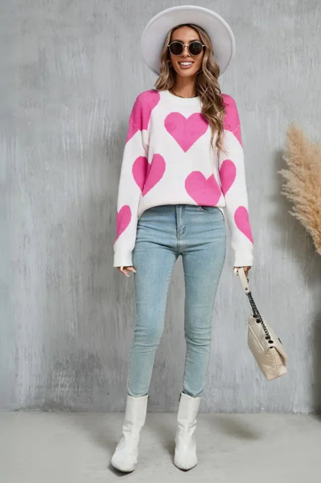 Woman wearing a white sweater with a pink heart print, light blue jeans, and white boots.