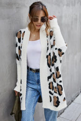 Woman leaning against a wall wearing a white leopard print long cardigan over a white tank top and blue jeans.