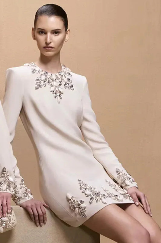 Model wearing a cream-colored, long-sleeved shift dress with elaborate crystal and pearl embellishments on the neckline, cuffs, and hem.
