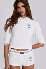 Front view of a model wearing a white cropped polo shirt with blue trim and matching white shorts with blue trim, displaying embroidered logos.