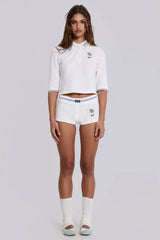 Full body shot of a model wearing a white cropped polo shirt, white shorts, and white socks with light blue ballet flats.