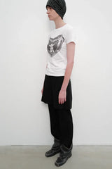 Side view of a model wearing a white crew neck t-shirt with a black and white graphic print on the chest, black shorts over black pants, and black boots.