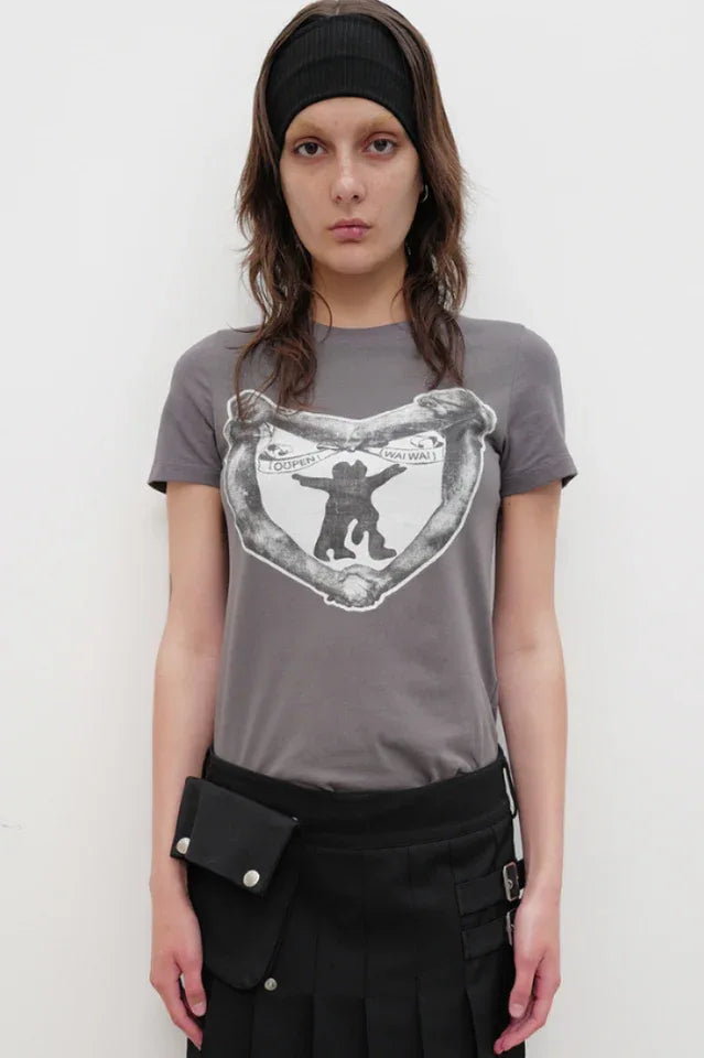 Model wearing a grey crew neck t-shirt with a black and white graphic print on the chest, a black belt bag, and a black pleated skirt.