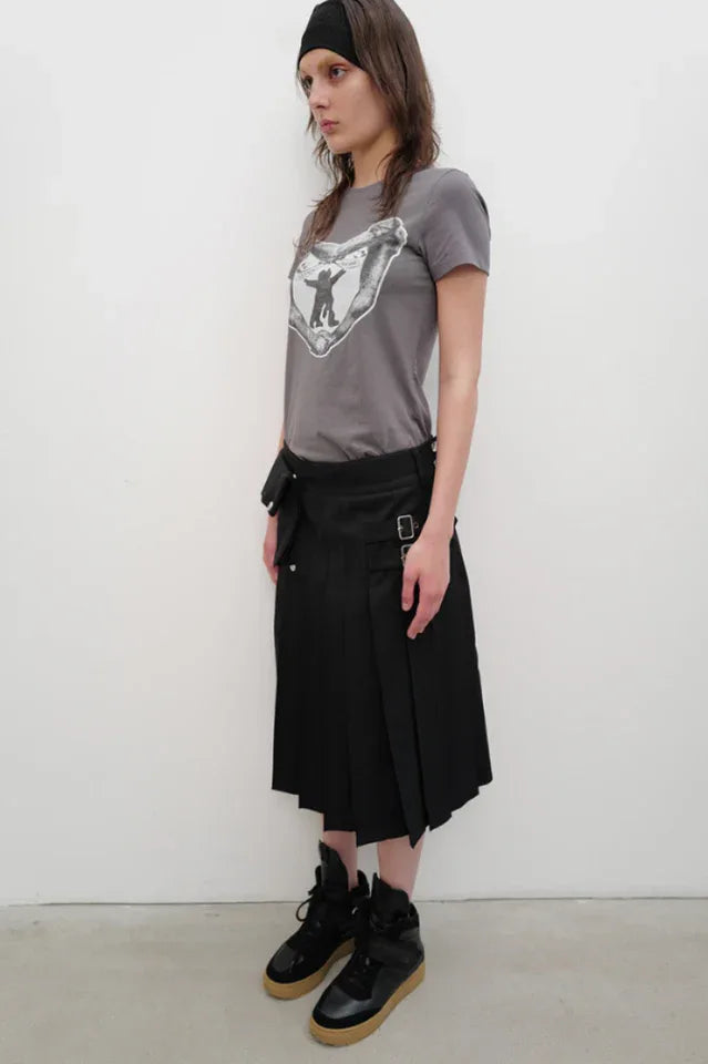 Side view of a model wearing a grey crew neck t-shirt with a black and white graphic print on the chest, a black belt bag, and a black pleated skirt.
