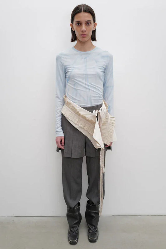 Model wearing a light blue tie-dye long-sleeve top, grey pleated trousers, and a beige draped overlay accessory.