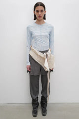 Model wearing a light blue tie-dye long-sleeve top, grey pleated trousers, and a beige draped overlay accessory.