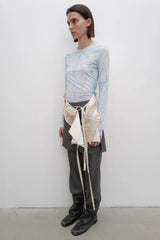 Side view of a model wearing a light blue tie-dye long-sleeve top, grey pleated trousers, and a beige draped overlay accessory.