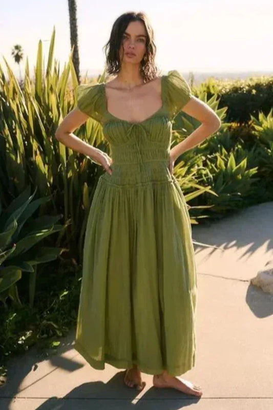 Woman wearing a green puff-sleeve midi dress with a smocked bodice and flowing skirt, standing outdoors with lush greenery in the background.