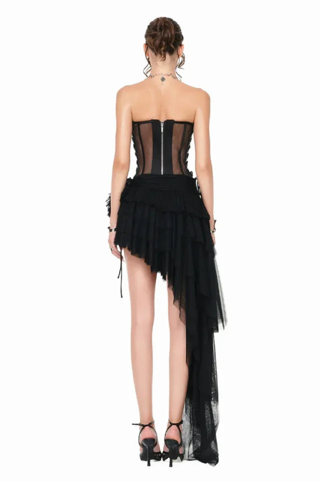 Woman wearing a black strapless corset dress with an asymmetrical ruffled skirt and long tulle train, back view.