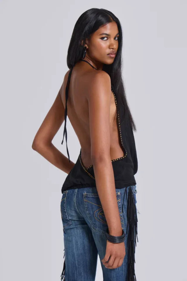 Back view of a model wearing a black halter top with an open back and studded trim, paired with blue jeans with fringe detailing.