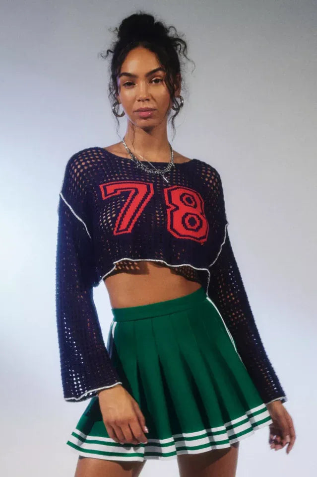 Woman wearing a navy blue cropped open-knit sweater with red appliquéd numbers and white trim, paired with a green pleated skirt.