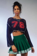 Woman wearing a navy blue cropped open-knit sweater with red appliquéd numbers and white trim, paired with a green pleated skirt.