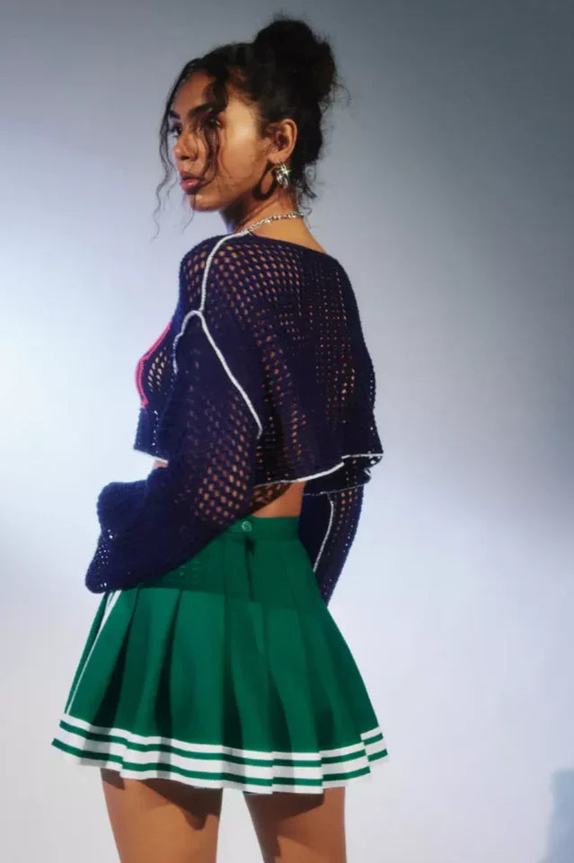 Back view of a woman wearing a navy blue cropped open-knit sweater with red appliquéd numbers and white trim, paired with a green pleated skirt.