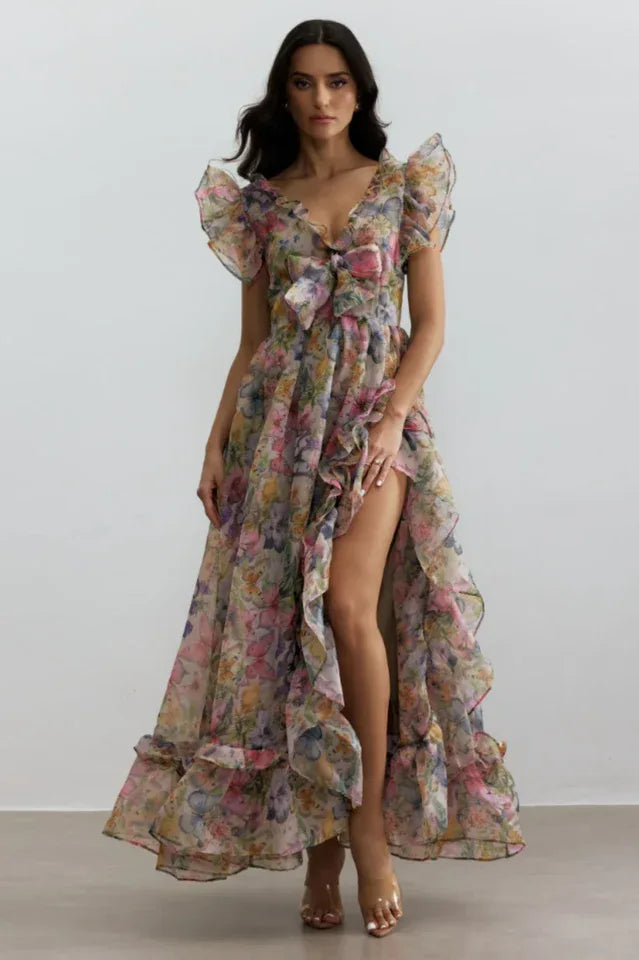 Woman wearing a floral print chiffon gown with V-neck, bow detail, flutter sleeves, and a high-low hemline with a thigh-high slit.
