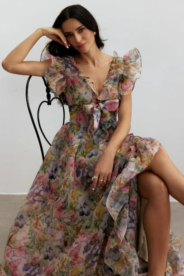 Woman seated, wearing a floral print chiffon gown with V-neck and ruffled details.