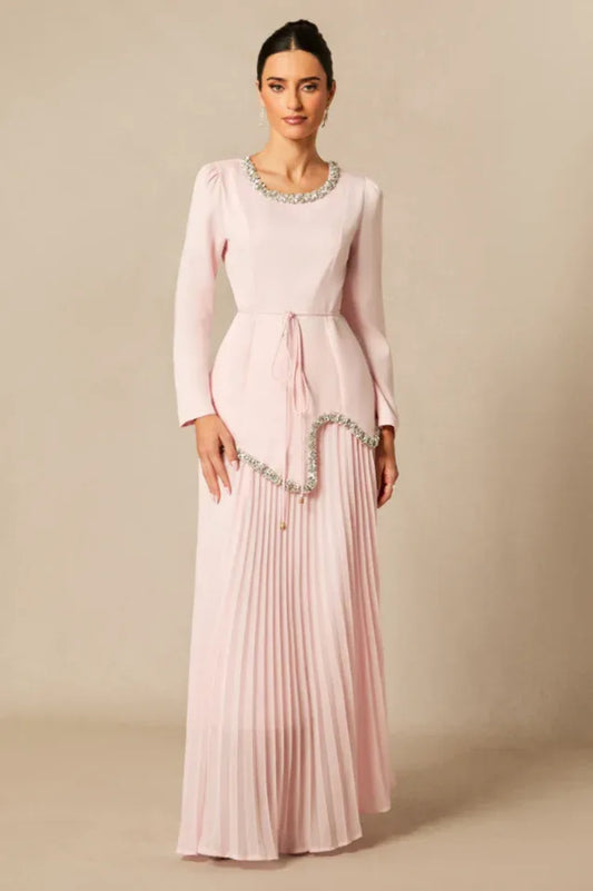 Woman wearing a long, light pink, long-sleeved dress with a round neckline and embellished trim, cinched waist with a tie, and a pleated skirt.