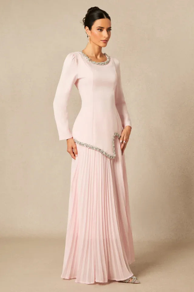 Side view of a woman in a long, light pink, long-sleeved dress with a round neckline and embellished trim, and a pleated skirt.