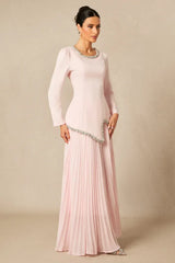 Side view of a woman in a long, light pink, long-sleeved dress with a round neckline and embellished trim, and a pleated skirt.