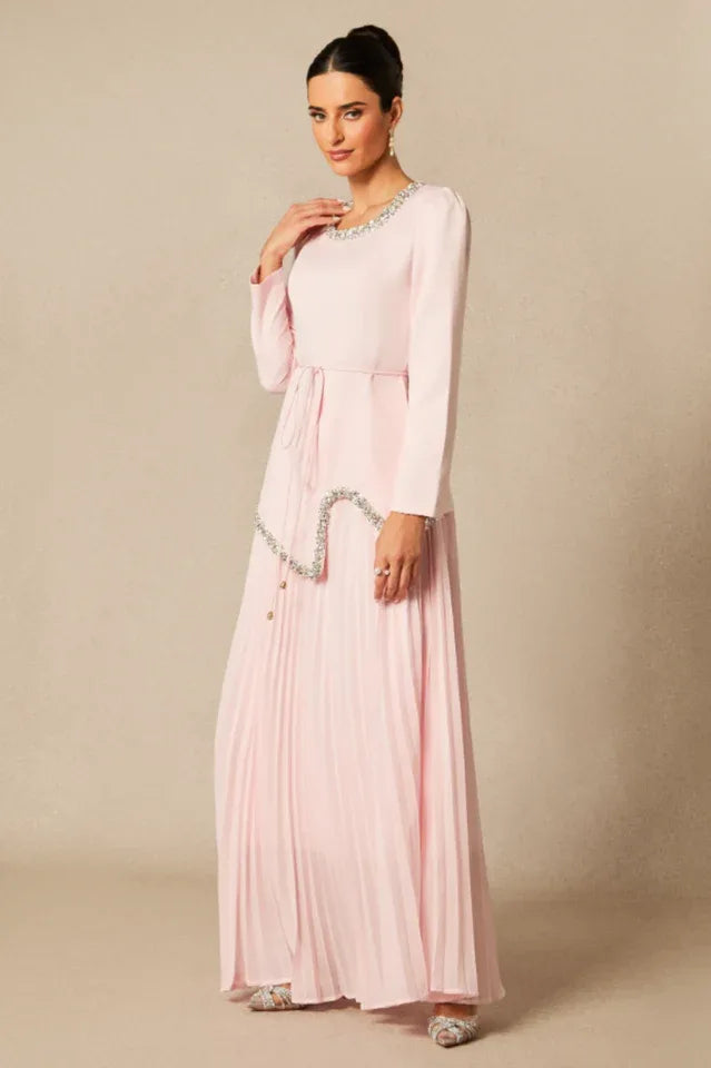 Woman posing in a light pink, long-sleeved formal dress with a pleated skirt and embellished details.