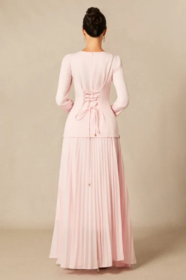 Back view of a woman wearing a light pink, long-sleeved formal dress with a pleated skirt and corset lacing detail.