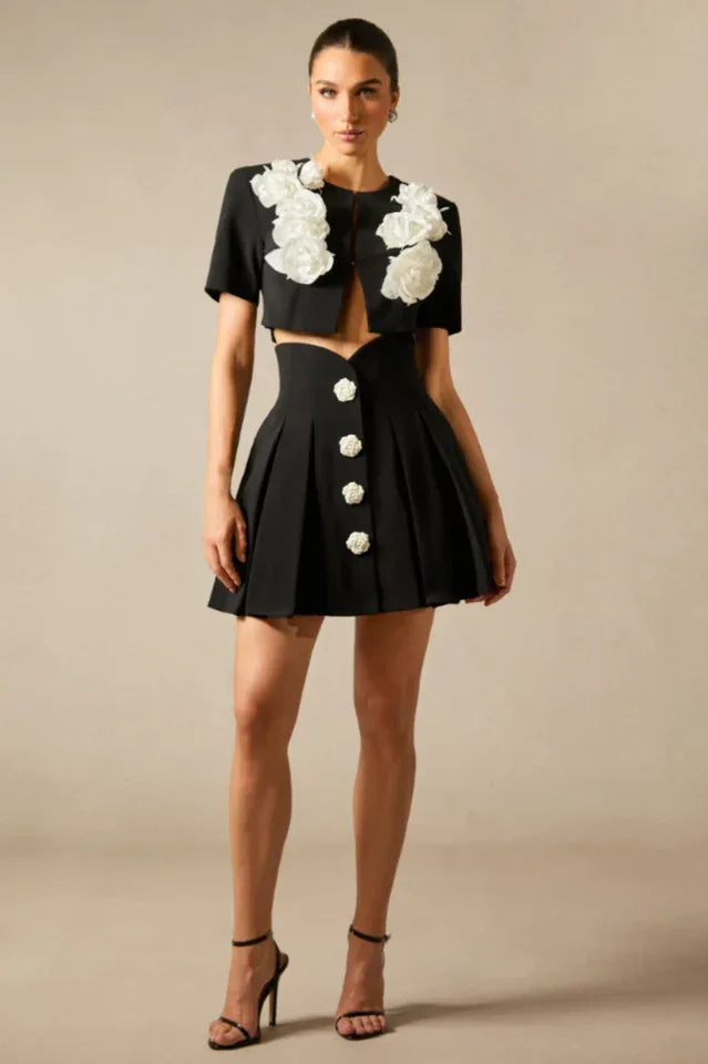 Full length view of a woman in a black cropped jacket and pleated mini skirt with floral accents.