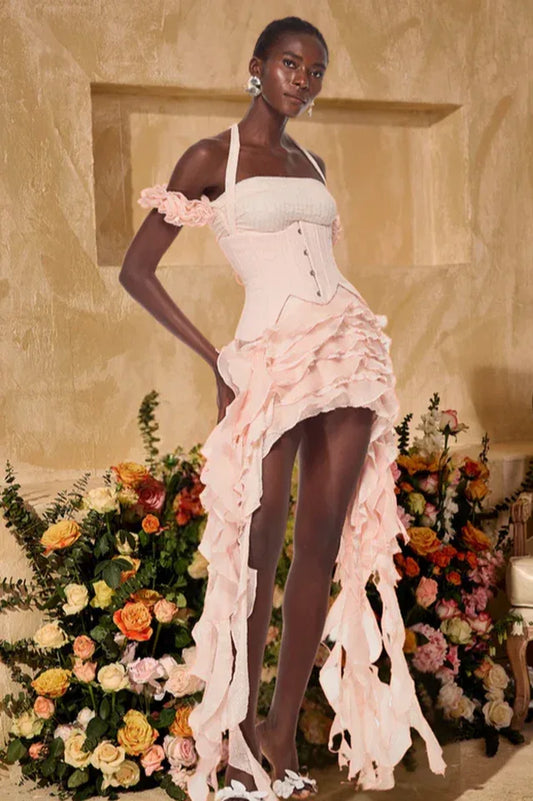 Model wearing a pink ruffled corset dress with a halter neckline and high-low hemline, standing in front of a floral arrangement.