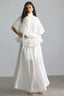 Woman wearing a white, flowing, high-neck dress with voluminous sleeves and a tiered ruffled waist.