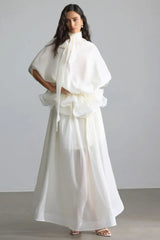 Woman wearing a white, flowing, high-neck dress with voluminous sleeves and a tiered ruffled waist.