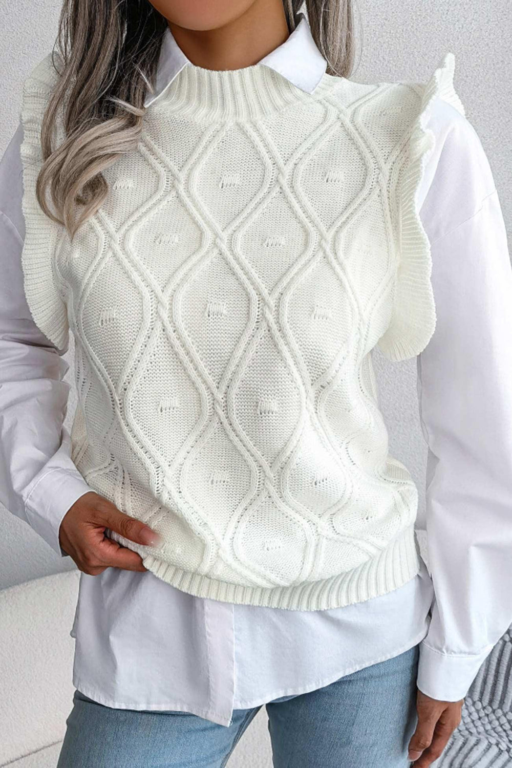 Woman wearing a white cable knit sweater vest with ruffled shoulders over a white collared shirt.