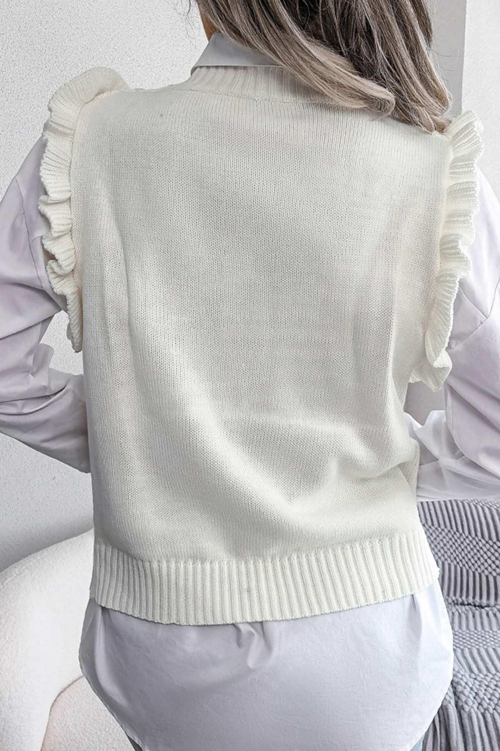 Back view of a woman wearing a white cable knit sweater vest with ruffled shoulders over a white collared shirt.