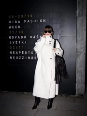 Woman in a white oversized wool blend coat standing in front of a black wall with event details