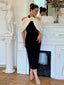 Woman wearing a black sheath dress with a white sheer off-the-shoulder overlay adorned with a large flower accent, standing in a formal setting.